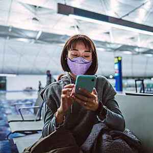 Young woman wearing a protective face mask taps on her smartphone as she waits for a night flight at a quiet airport terminal gate.